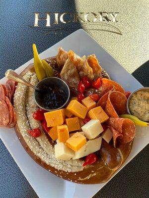 A Pretzel Charcuterie Plater with diff ent cheeses, meats, and picked vegetables.