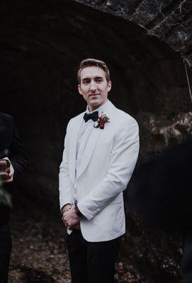 Groom in white on white tux with black pants stands waiting