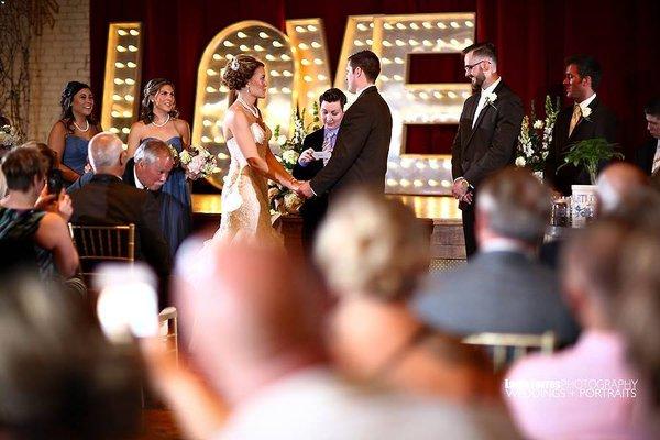 A bride and groom exchanging vows saying I do with our lighted love marquee letters in the background