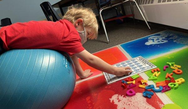 Child working on core strength while prone on yoga ball and reaching for letters