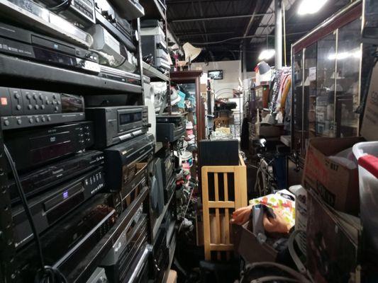 An extremely narrow path showing a shelf full of random stereos and more.