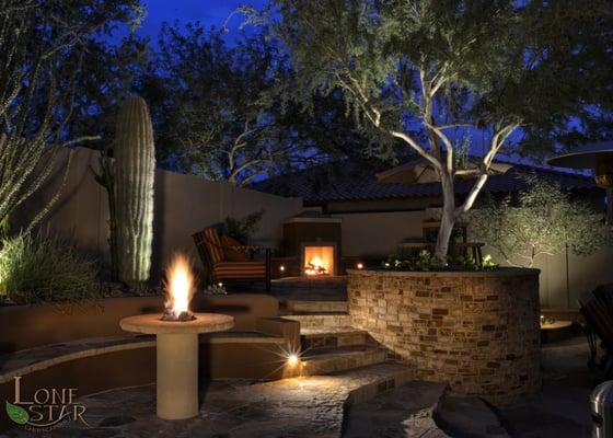 This backyard features a fire table, bench, and fireplace all highlighted with low voltage landscape lighting.