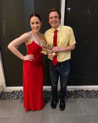 A white woman in a red dress and a white man in a yellow shirt, red tie, and jeans holding awards and smiling.