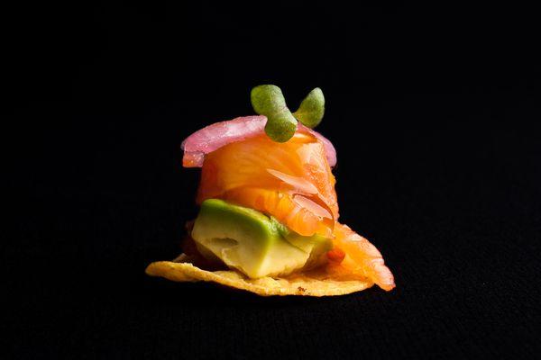 Salmon Tostadas are served on a corn tortilla with house cured salmon, avocado, aji-ponzu, picked red onion & micro greens