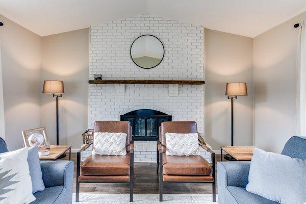 living room with fireplace, white brick, wood mantle, vaulted ceiling, vinyl plank flooring, round mirror, brown chairs, grey couches