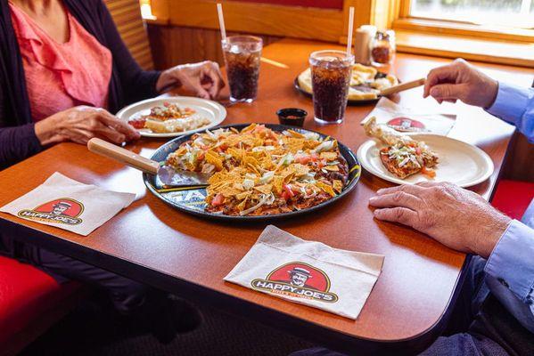 A couple sitting around a table at a Happy Joe's restaurant with a Taco Joe Pizza between them.