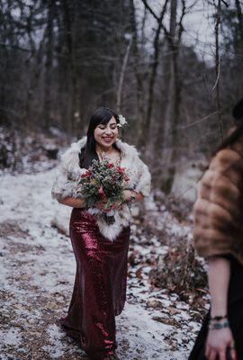 Bride in fur shawl and red dress walks down trail, holding a greenery and red lilies bouquet