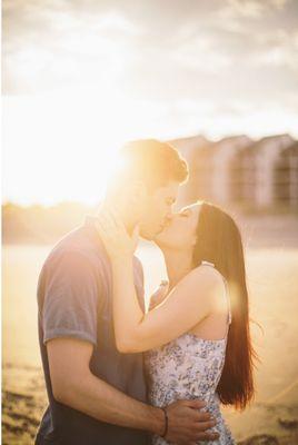 Newly engaged couple, bathed in warm golden sun, kiss on the beach.