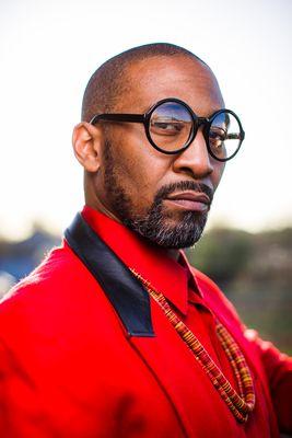 Portrait of a man with black glasses and red suit with beaded necklace.