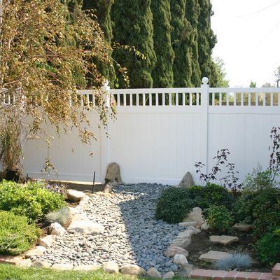 A white vinyl privacy fence is the center of the photo. With trees in the background and some rocks and plants in the front of the fence.