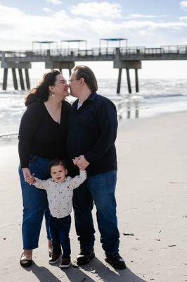 Mom and Dad loving on each other while holding onto son at Jacksonville Beach Pier near Jacksonville, FL