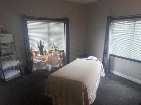 Relax in our tranquil massage room featuring lush plants, a plush table, warm towels & serene silence for ultimate pampering.