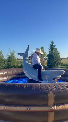 A man in a white button down shirt and a beige cowboy hat, rides a large mechanical shark surrounded by a brown and blue inflatable ring.