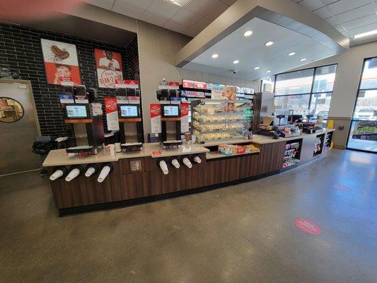 3 Coffee dispensers are here. Doughnuts are in the display case to the right of the coffee dispensers. Cash register to the right.