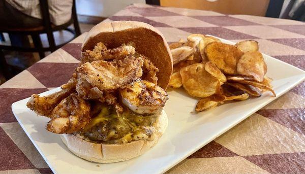 Surf and turf burger with blackened shrimp, cheese, fried onion strings, and garlic sauce. Homemade potato chips on the side