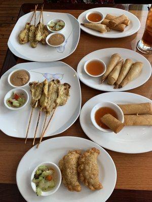 2 plates of appetizer chicken sate, 2 orders of appetizer cream cheese rolls, funnel shaped shrimp rolls and chicken curry puffs in front.