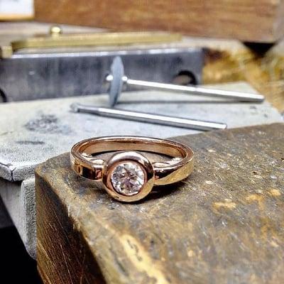 A bezel-set diamond ring in rose gold is on the bench for finishing.