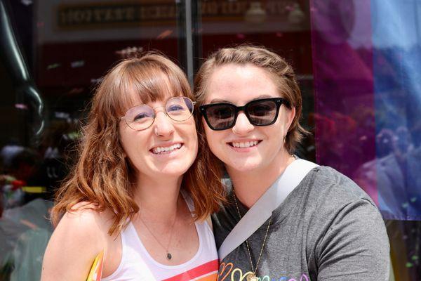 Two people smiling with a pride flag in the background.