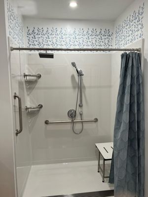 Wheelchair accessible showers for the whole family, with trench drain, personal shower and rain head. Dual purpose grab bars/ corner shelves