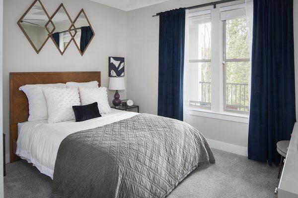 Elegantly staged bedroom with a wooden headboard, plush bedding, geometric mirrors, deep blue curtains, and subtle art accents.