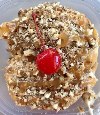 A cinnamon roll with maple cream cheese frosting, pecans, caramel, and a cherry.