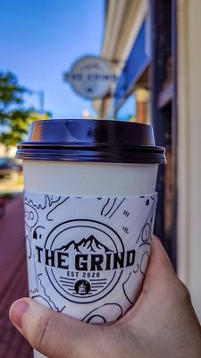 Holding a to-go cup from The Grind, with my hand in front of me, and the building sign for the coffee shop out of focus in the background