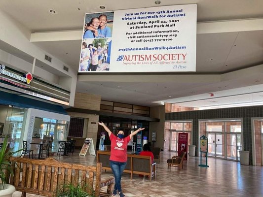 Our President, Aime Phillips, stands underneath a banner displaying the 13th Annual Run Walk for Autism at Sunland Park Mall