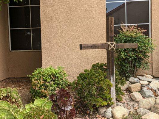 Beautiful wooden cross in a bed of rocks and shrubs, windows in the background