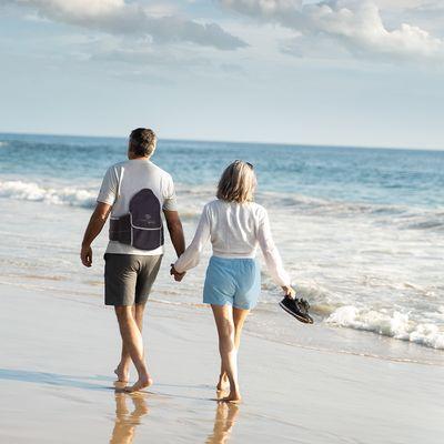 A couple walks along the shore, holding hands. The man strides confidently, supported by the TRUverse LSO Back Brace, enjoying every moment.