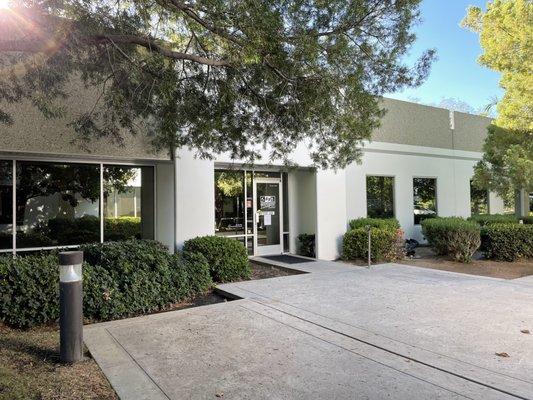 ILCKC Front door image from left side of main entrance. Trees and bushes aside concrete walkway with pushbutton for door on the right side.