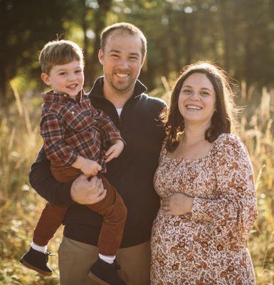 Family photo of pregnant woman, husband and toddler in fall clothes, standing outdoors in a field of tall grass and golden sun.