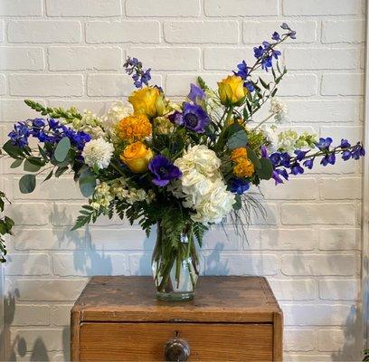 Sympathy arrangement stands approximately one and a half feet tall and includes yellow roses, delphiniums, Pom Pom Ranunculus and anemones.