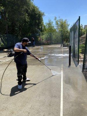 Ivan is pressure washing a concrete tennis court.