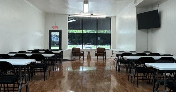 Spacious front view of our event space with stylish tables, chairs, a modern TV, and privacy-filmed windows