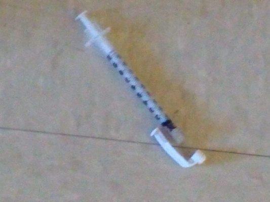 Syringe on floor next to garbage pail