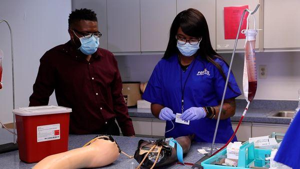 Medical Assisting instructor watching a student prepare needle for blood draw on dummy arm