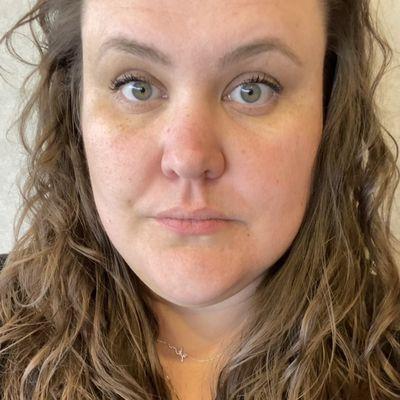 Face of a white femme with brown curly hair. The hair is damaged and the curl pattern is uneven.