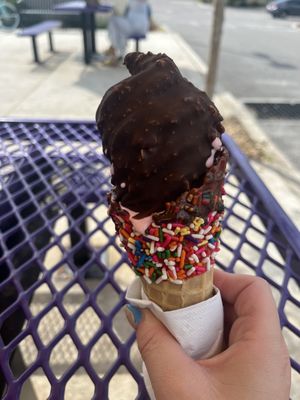 Strawberry soft serve ice cream dipped in chocolate with a waffle rainbow sprinkled cone.