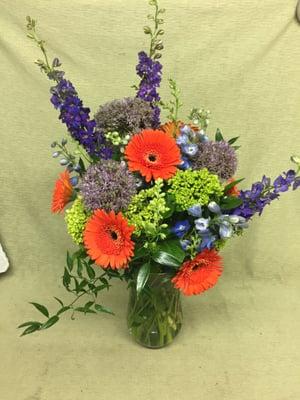 Orange gerber daisies, purple larkspur, green mini hydrangeas, purple allium, blue delphinium, and greenery in a clear glass vase.