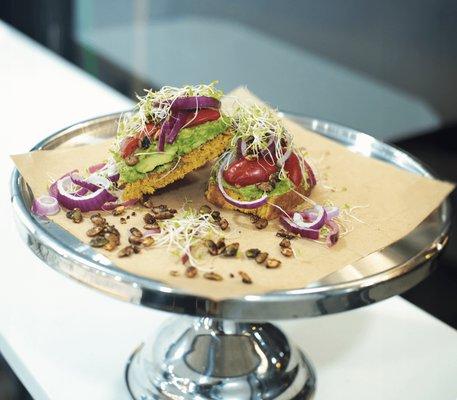 Avocado Toast -Fresh avocados on vegan coconut turmeric bread topped with sprouts, red onions, cherry tomatoes and toasted pumpkin seeds