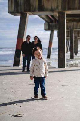 Toddler standing underneath the Jacksonville Beach Pier with parents in the background near Jacksonville, FL