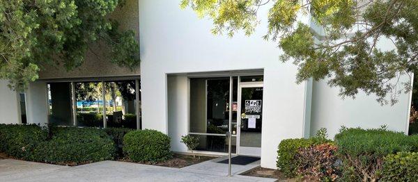 Front door from left side. Concrete walkway leads to door with a pushbutton extended up from a poll on the right side of the door walkway.